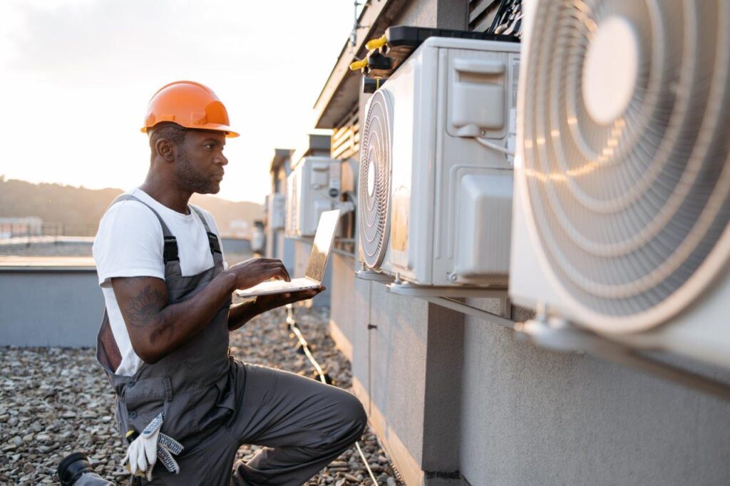 man typing on wireless laptop while kneeling near hanging air conditioner outdoors