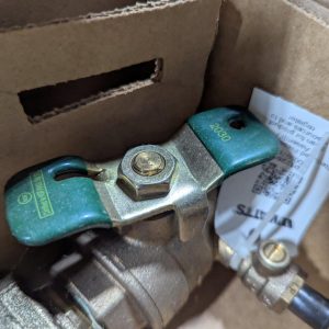 Close-up of a green metal valve handle with a central hex bolt, mounted on a pipe inside a cardboard box, with a tag tucked beside it.
