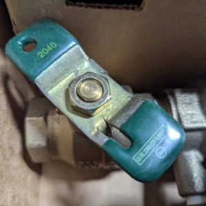 Close-up of a green metal valve clamp with a central hex bolt on a brown pipe surface; appears to be a shut-off or throttle valve component.