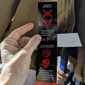 Slim black garment tag held by a hand, reading 'Stretch Durability' with red icons and notes on wind and water resistance.