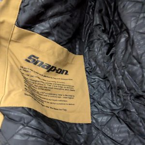 Close-up of a tan Snap-on fabric label sewn onto a black quilted jacket, with small printed text visible on the label.